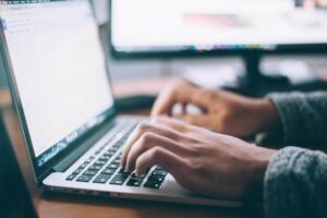 Eine Person mit einem grauen Pullover Tippt mit der Tastatur an einem Macbook etwas ein. Es soll das KI Texte schreiben symbolisieren.