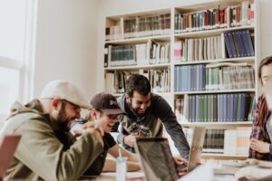 Mehrere Menschen stehen und sitzen an einem Tisch vor einem Bücherregal wir in einer Bibliothek und lachen.